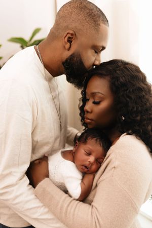Intimate family portrait in soft neutral tones showing parents tenderly embracing their sleeping baby in white. Dallas newborn photographer