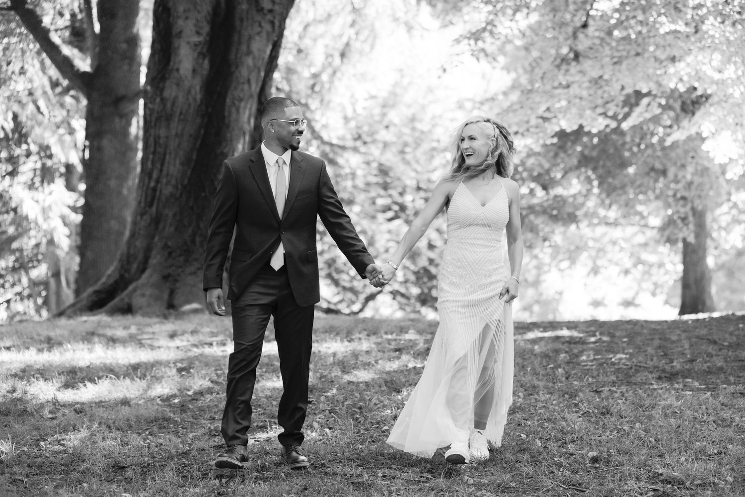 Black and white photo captures couple holding hands while walking through a majestic forest setting.