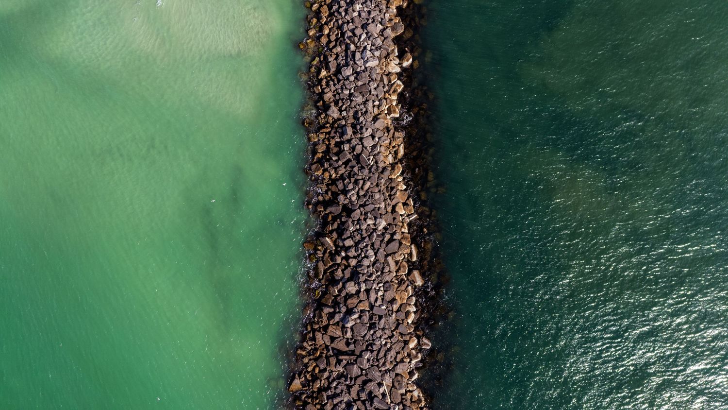 Aerial top-down view of a stone breakwater dividing turquoise green waters.