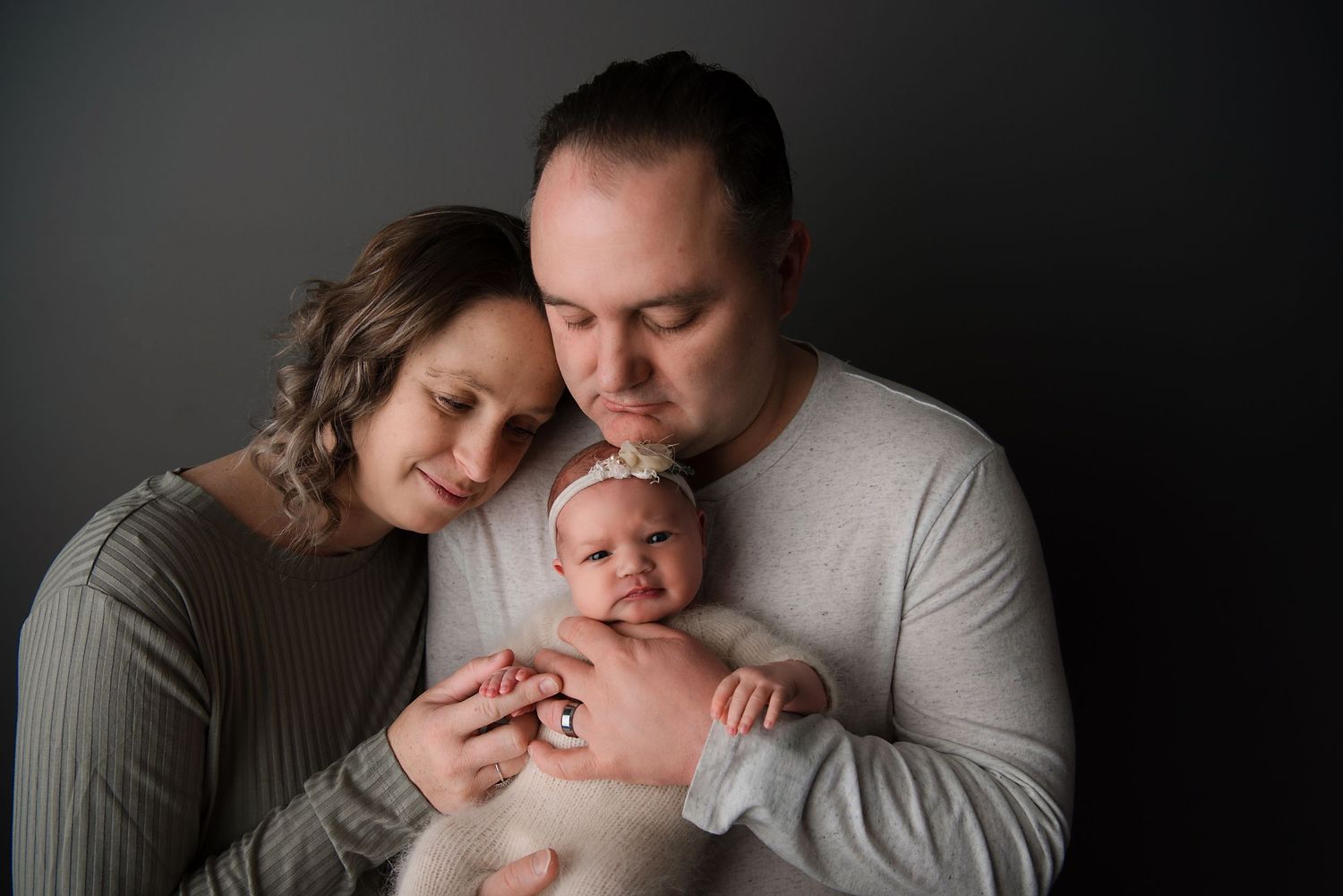 A couple in gray clothing snuggles close to their newborn baby in a moody studio portrait.