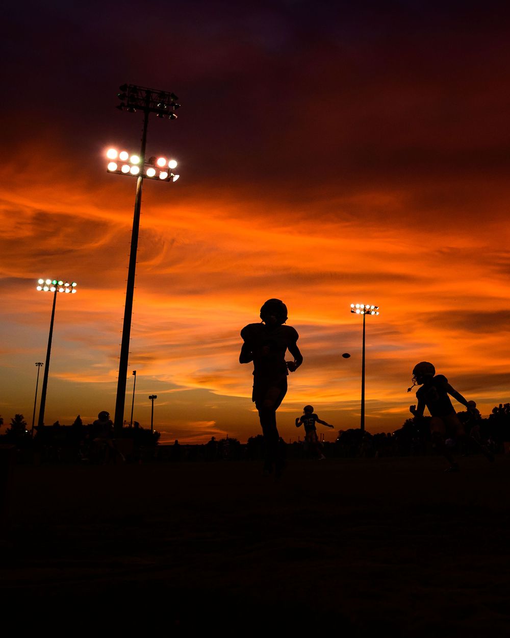 Silhouetted football players in helmets run across a field beneath stadium lights and a vivid orange and purple sunset sky.