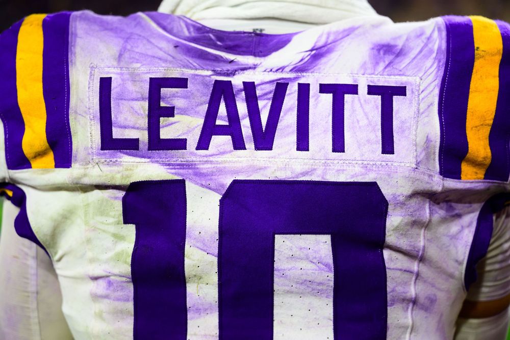 Back of a dirt-stained white football jersey with purple and gold shoulder stripes, showing the name Leavitt above the number 10.