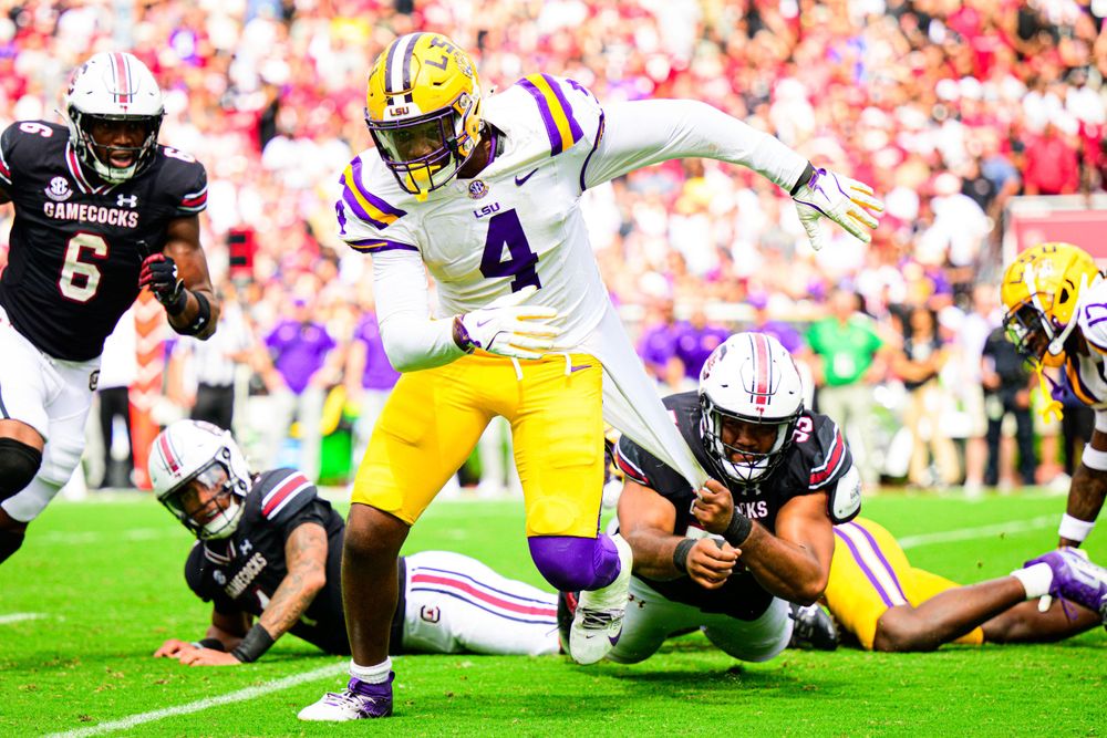 LSU football player jumps over defensive line during an intense game against opposing team at packed stadium.
