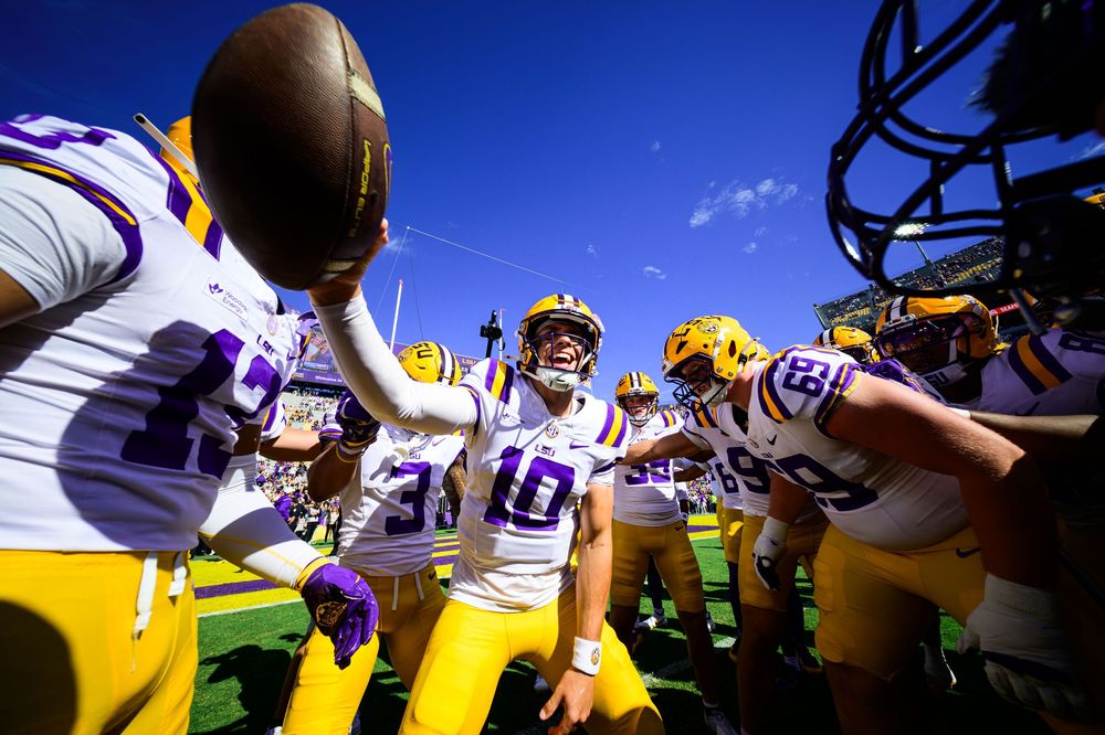 Football player in white number 10 jersey and gold helmet holds a football high as teammates huddle around him on a sunny field.