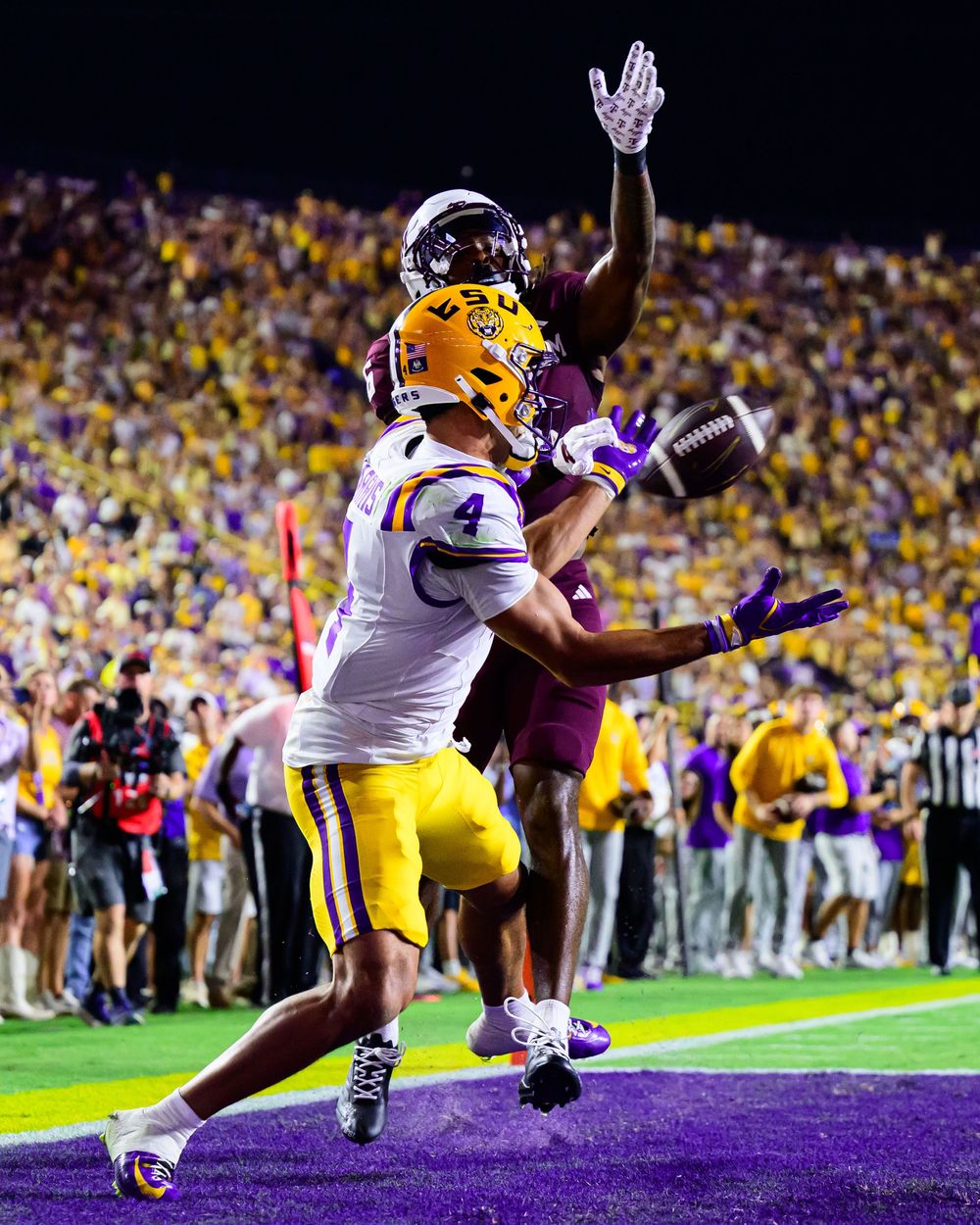 A LSU football receiver in a white and gold uniform leaps for a pass while a defender in a maroon jersey contests it in a packed stadium.