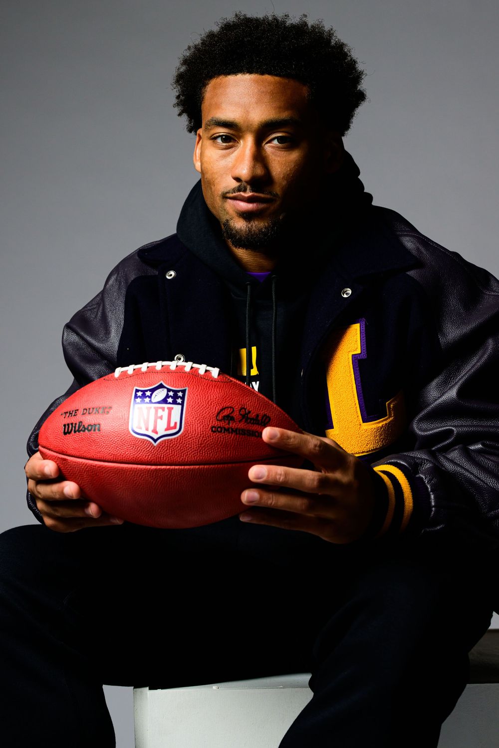 Professional athlete in varsity jacket poses with official NFL football against gray studio backdrop.