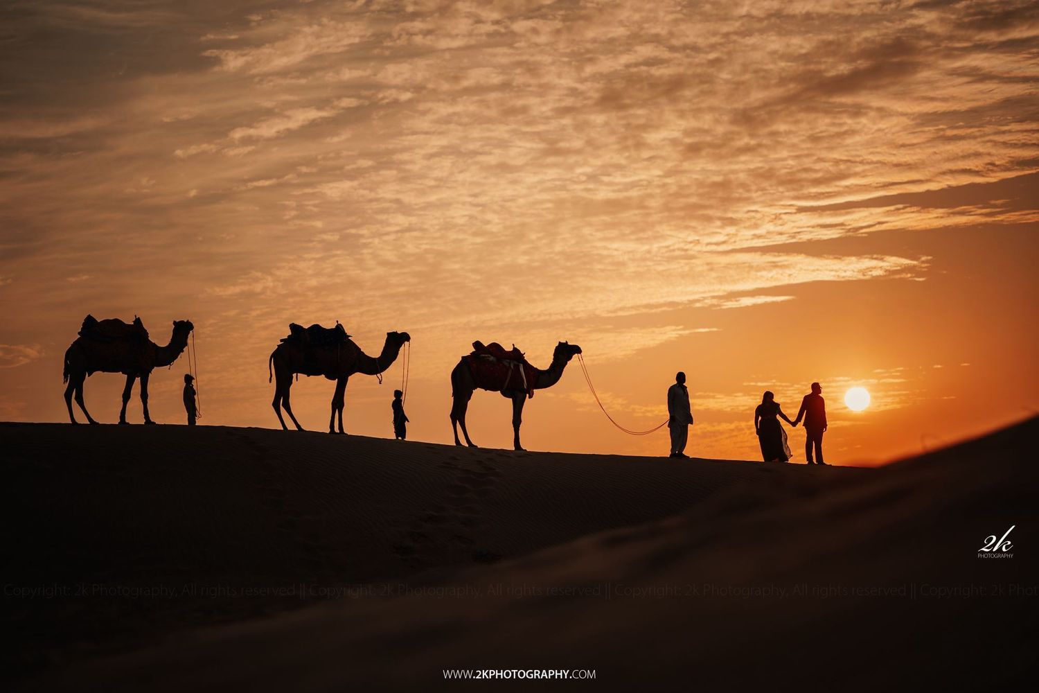 Silhouettes of three camels and people walking across desert sand dunes against dramatic orange sunset sky.
