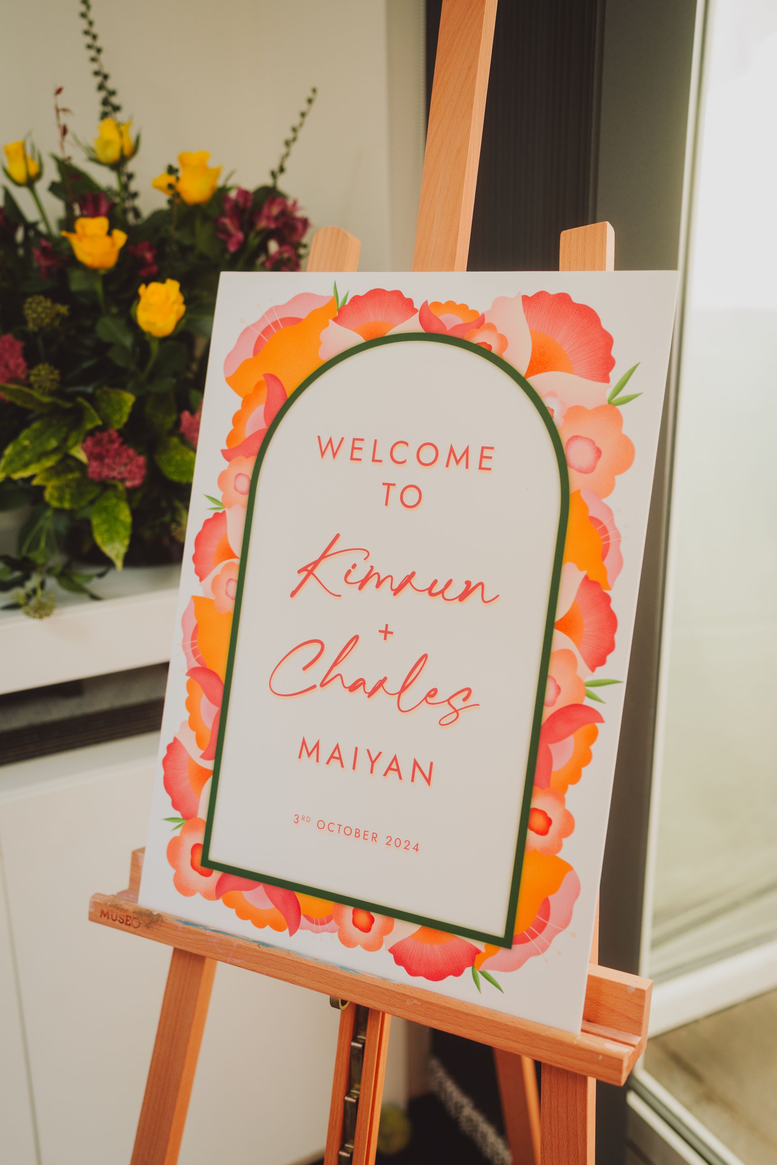 A floral welcome sign on an easel reads 'Welcome to Kimran + Charles Maiyan' with orange decorative elements.