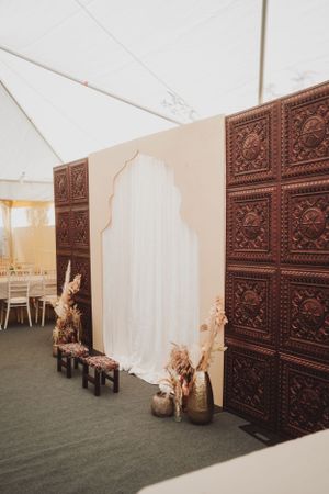 Decorative brown carved wooden panels create an elegant backdrop with white curtains and dried floral arrangements.