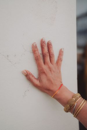 A hand adorned with mehndi designs and bangles rests against a white wall.