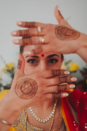 Hands adorned with henna designs and jewelry frame the decorated face of someone in traditional Indian attire.