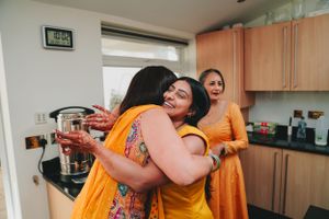 Friends sharing joyful hugs in a kitchen while wearing vibrant yellow and orange traditional Indian attire.