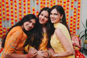 Three friends in colorful Indian outfits posing together against orange decorative backdrop.