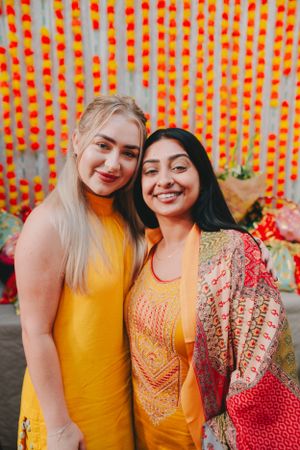 Two friends in yellow outfits smile together against orange floral backdrop.