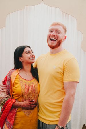 A happy couple embraces while wearing vibrant traditional Indian attire against a white archway background.
