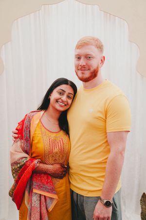 A couple poses together against a white backdrop wearing yellow and orange traditional Indian clothing.
