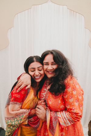 Two women in colorful Indian outfits share a joyful embrace while laughing together.