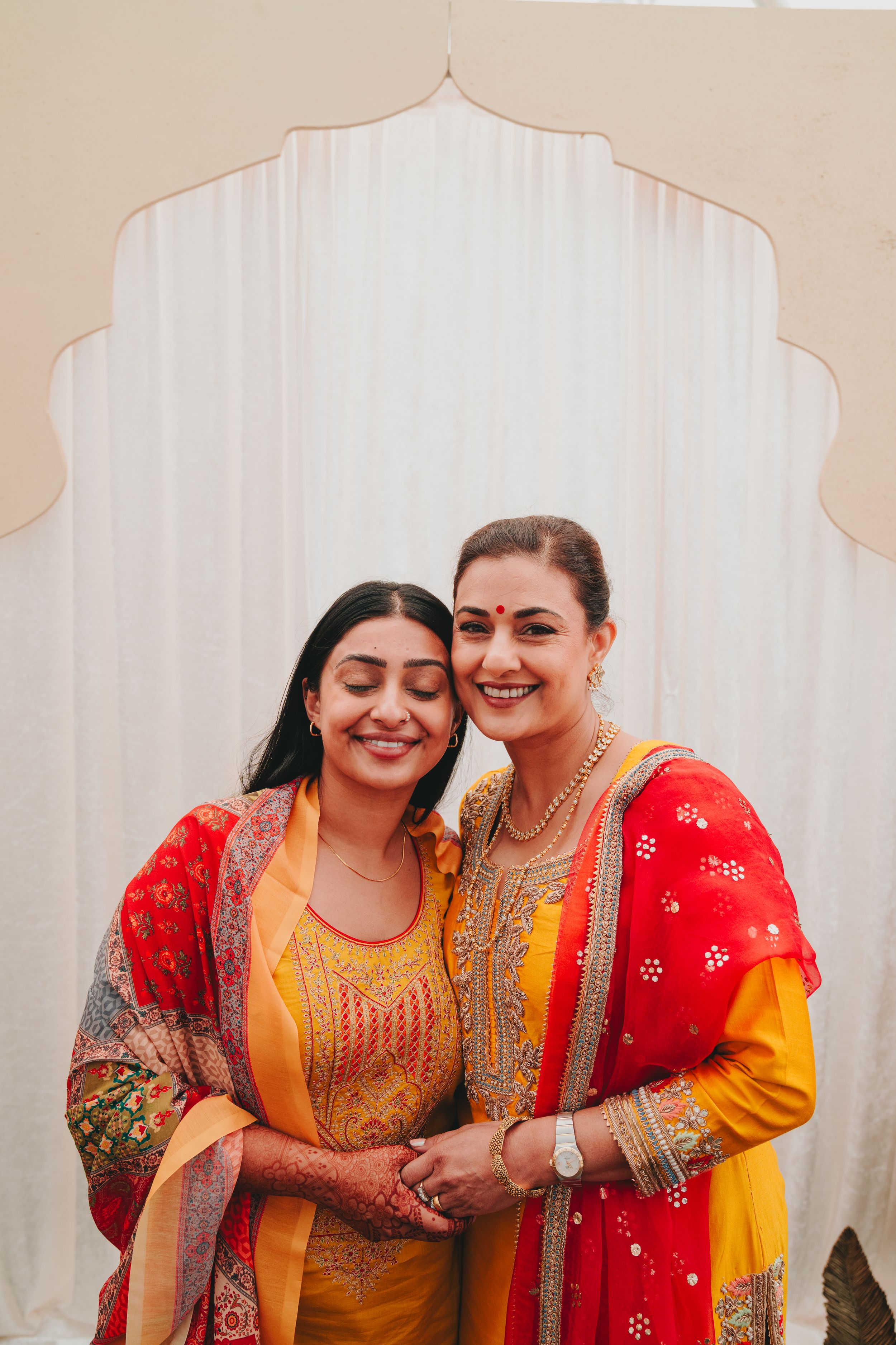 Two women in colorful traditional Indian attire share a joyful moment against an ornate backdrop.