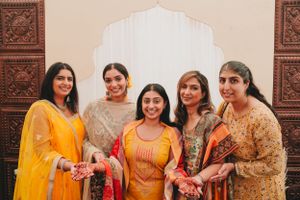 Five women in colorful traditional Indian outfits pose together against an ornate backdrop.