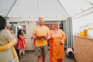 People in bright orange clothing carrying plates under a white tent at a cultural event.