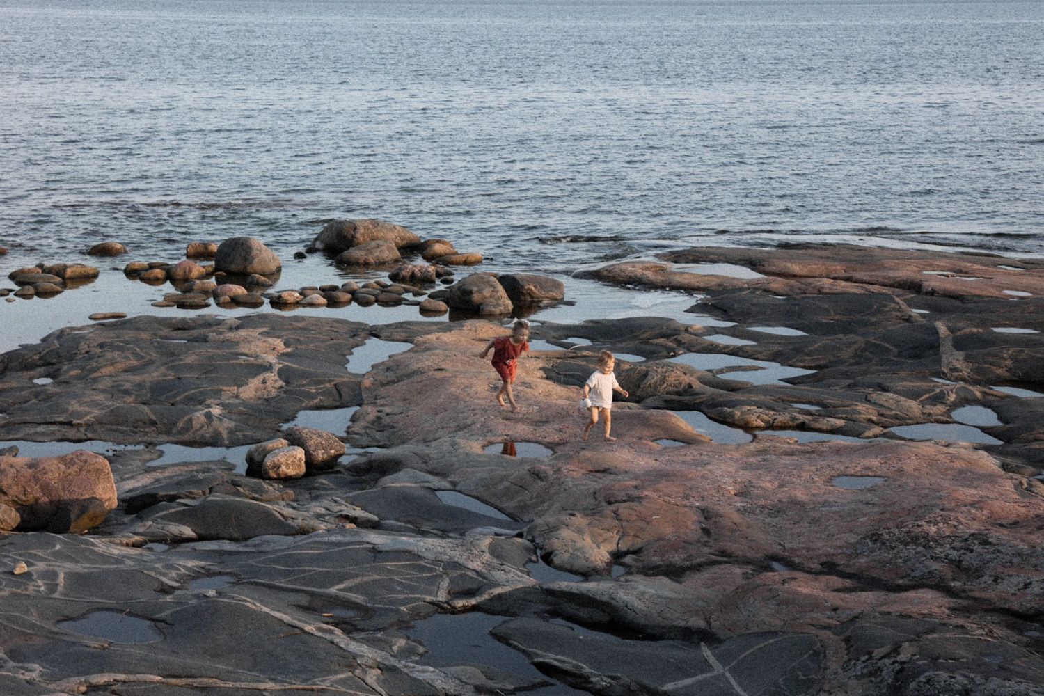 Kaksi lasta tutkii rantaviivaa auringonlaskun aikaan rauhallisilla rantakallioilla Inkoossa kesäiltana dokumentaarisessa perhekuvassa.
