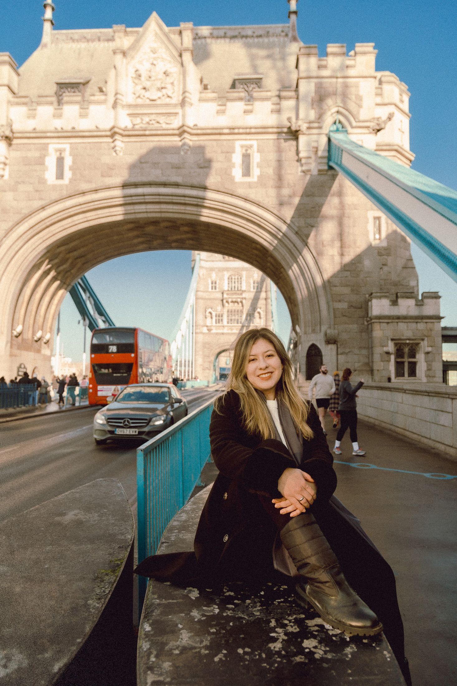 Tourist posing on Tower Bridge walkway with red London bus visible in background.