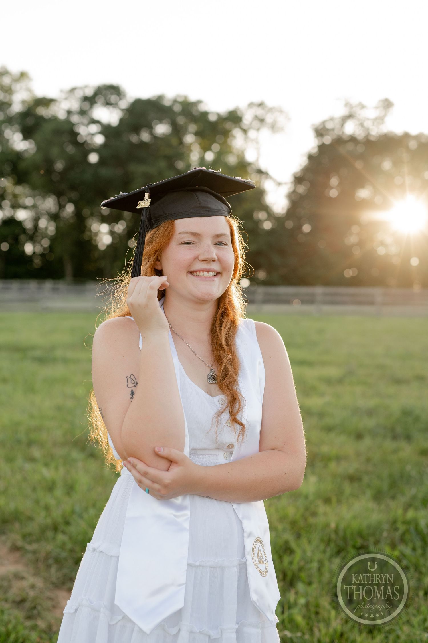 Graduate in cap and gown smiling at sunset in rural field setting.