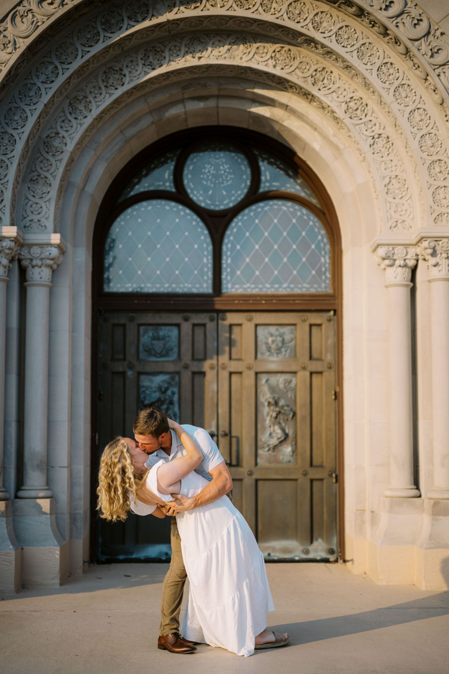 A romantic moment between a couple sharing an intimate dip kiss in front of an ornate church doorway with arched architecture.