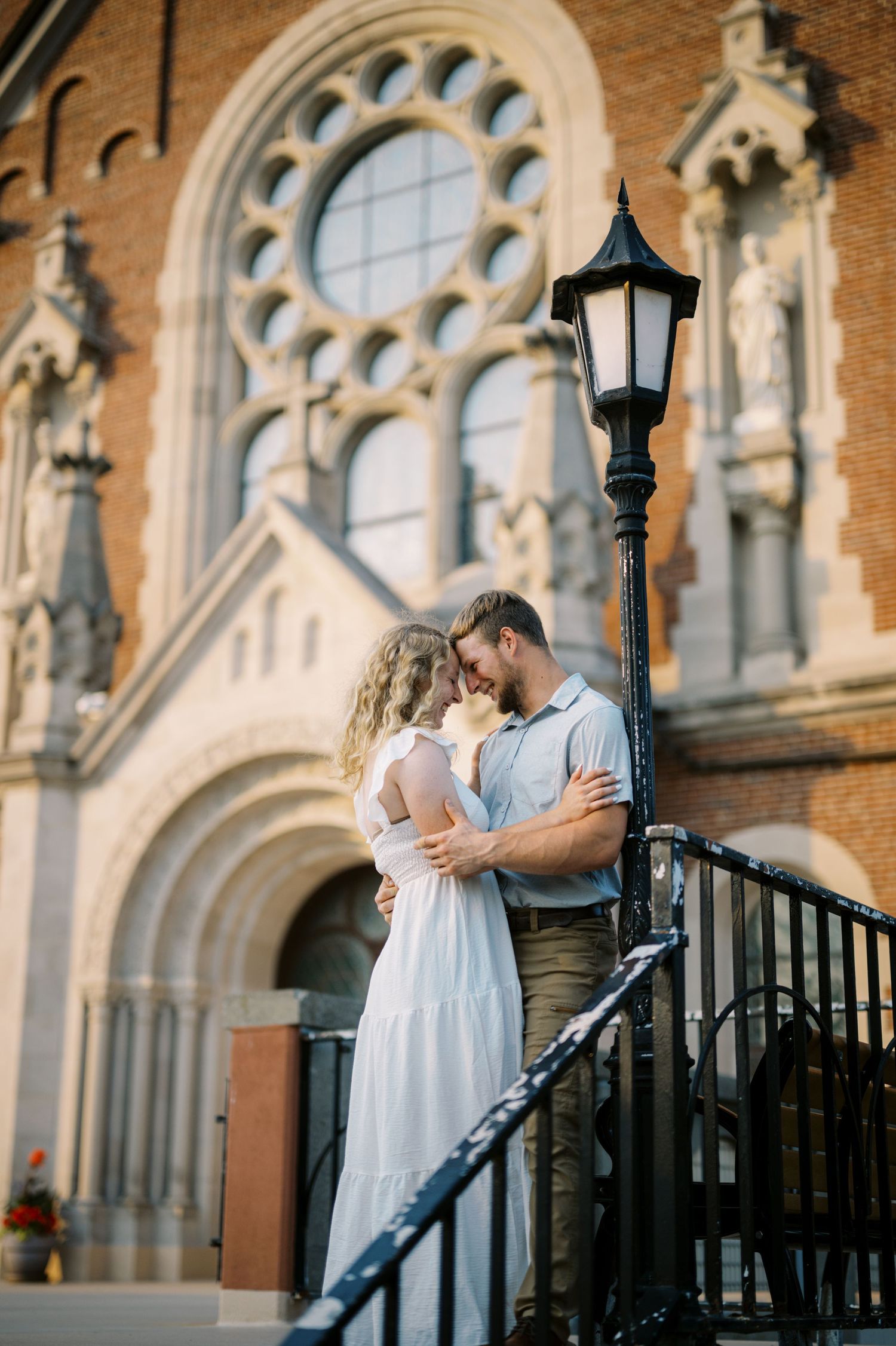 A romantic couple embrace on church steps with ornate Gothic architecture and a vintage lamppost in the background.