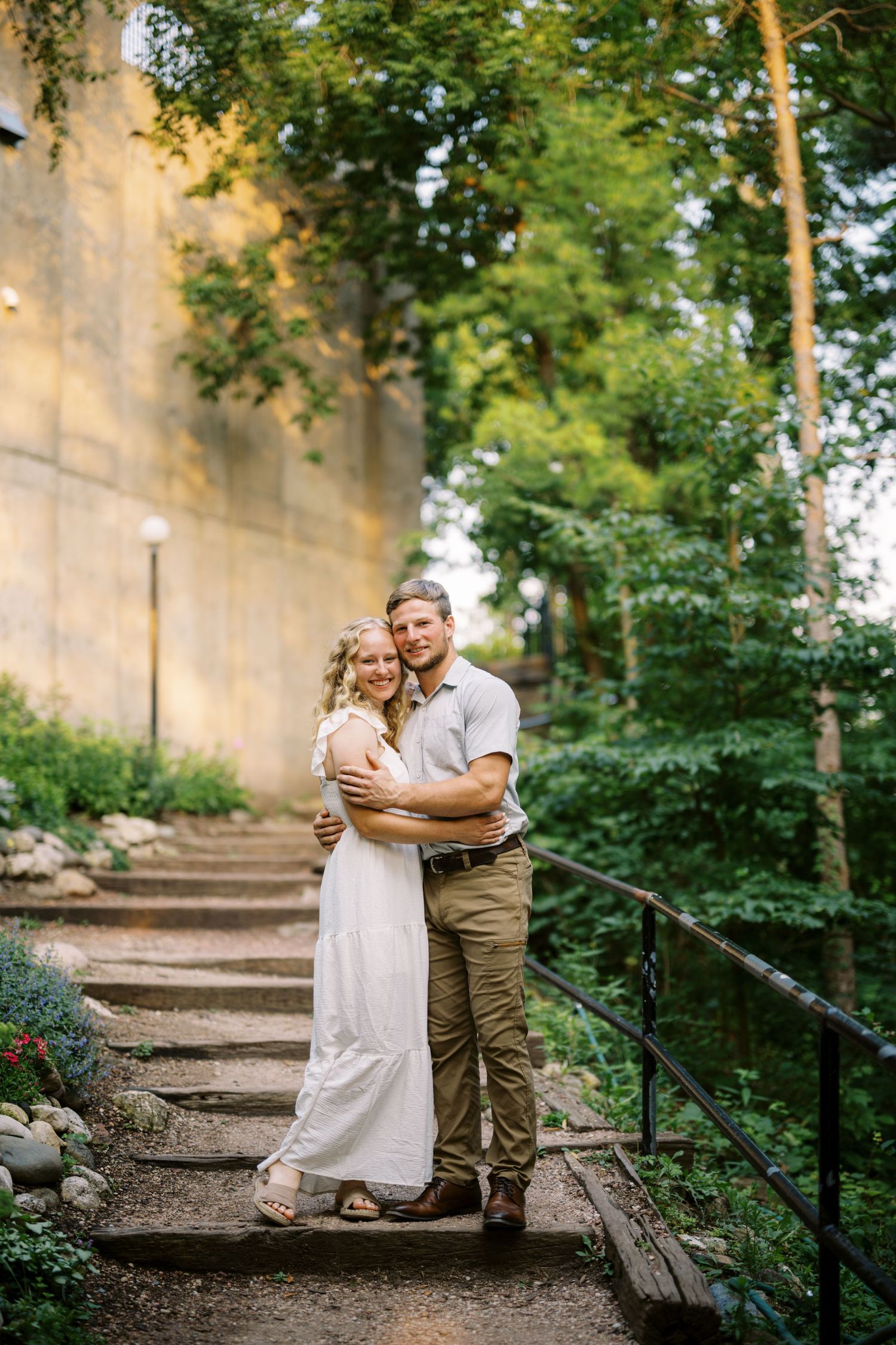 A couple embraces on rustic stone steps surrounded by lush greenery and trees on a sunny day.