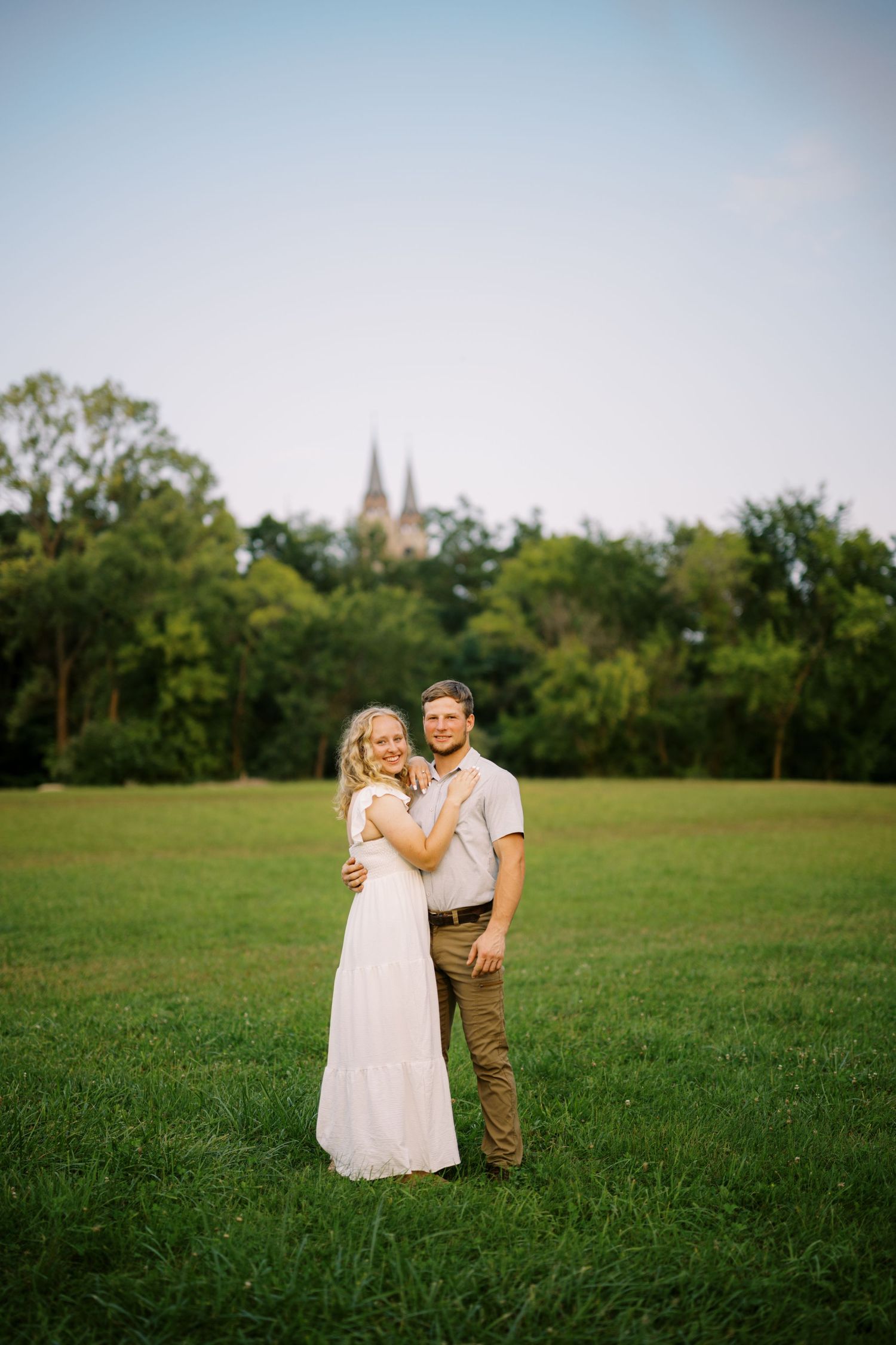 Romantic couple embraces in a green field with church spires visible in the background at sunset.