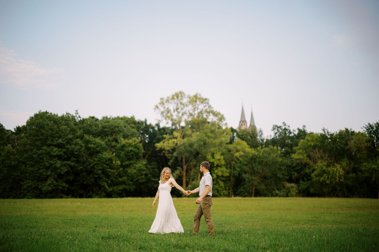 A couple holds hands while dancing together in a grassy field with church spires visible in the distance.