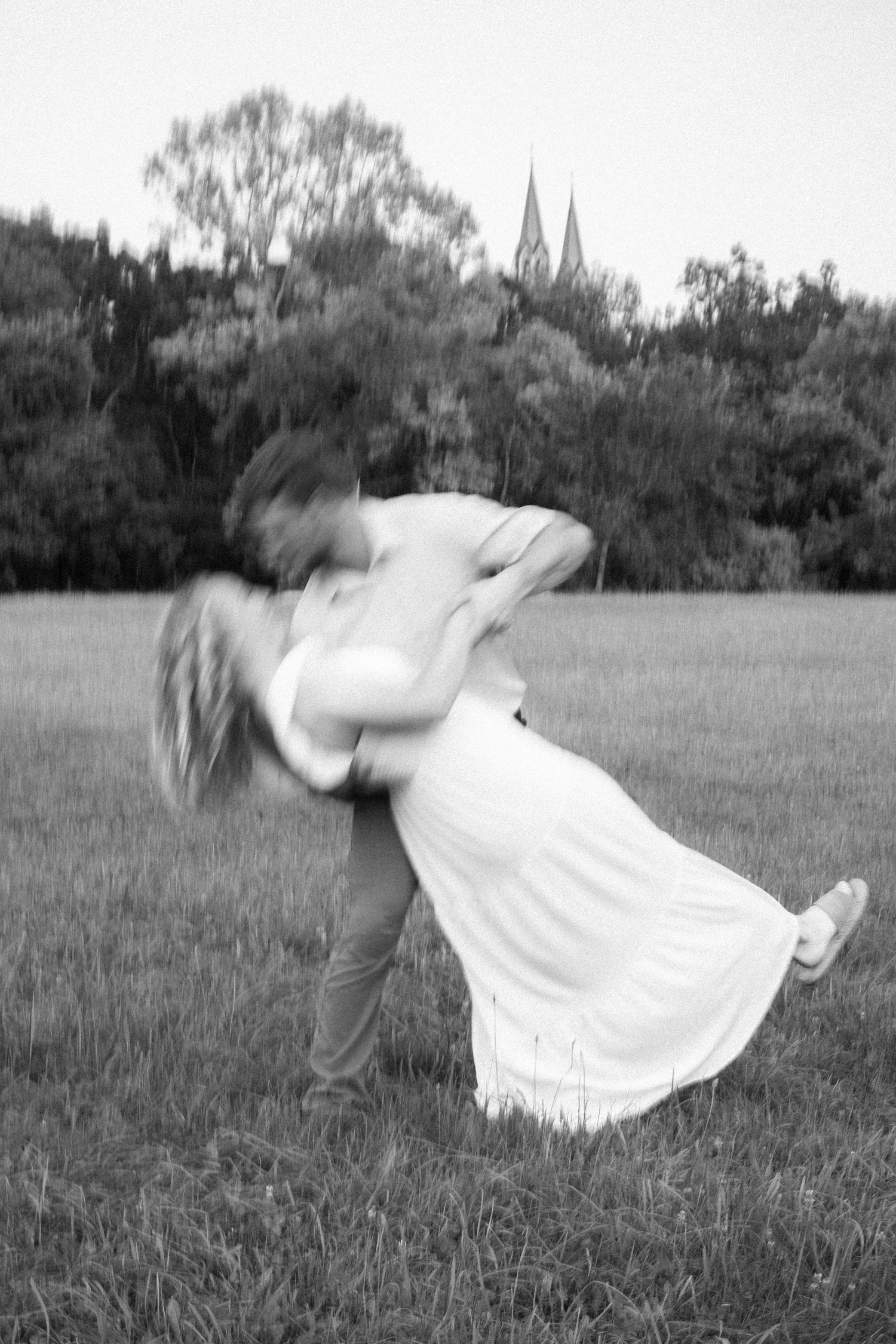 A black and white photograph of a couple dancing in a field with church spires visible in the distance.