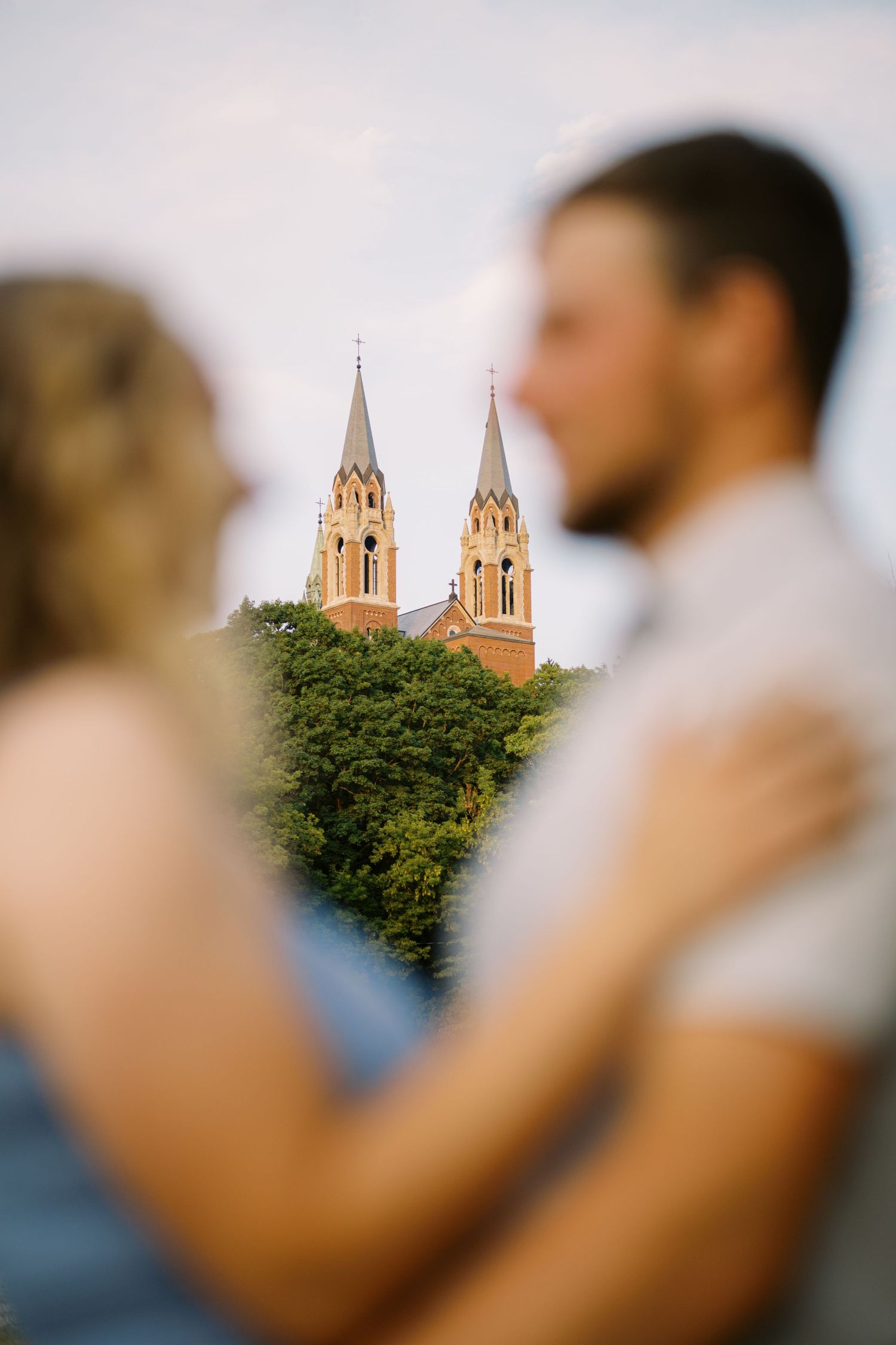 Church spires rise above trees in soft focus as a couple embraces in the blurred foreground.