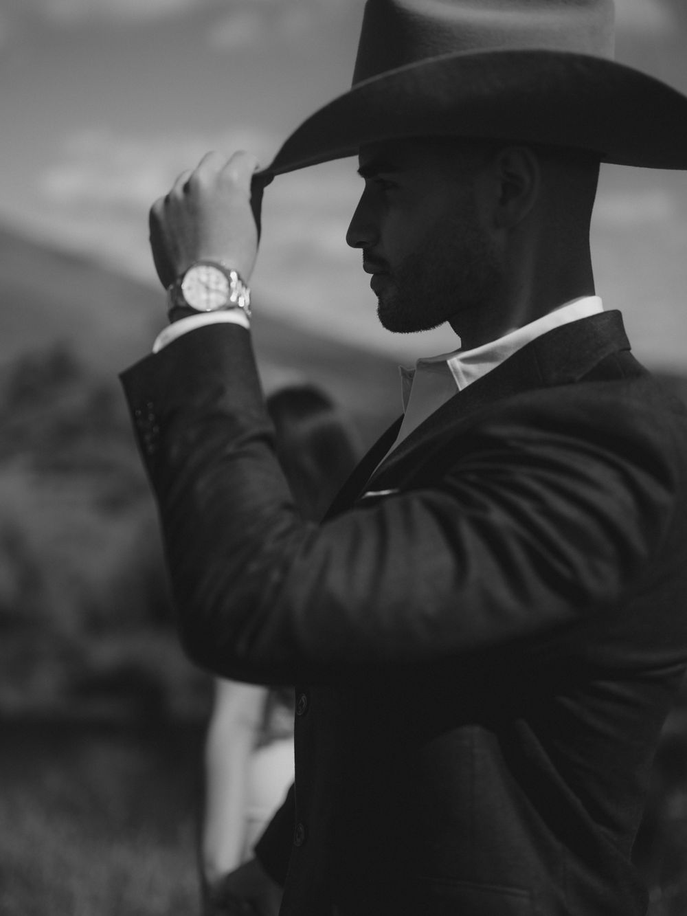 Figure in formal suit and cowboy hat adjusts brim against mountainous backdrop.
