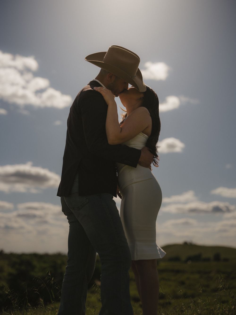 Romantic couple embracing in western attire against cloudy countryside backdrop.