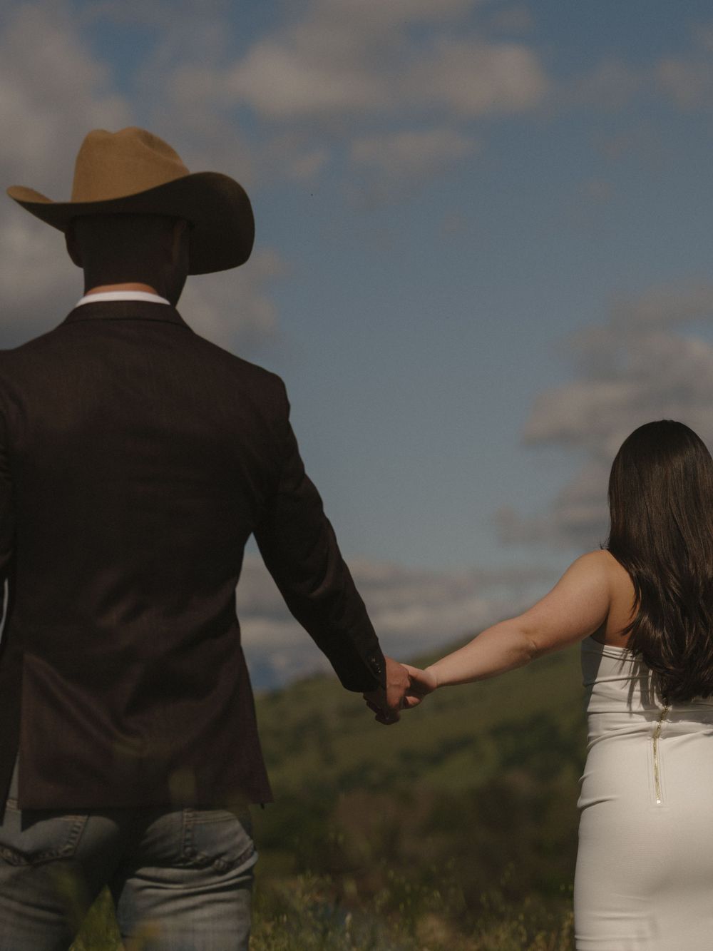 A couple in formal attire holds hands while walking through a scenic hillside landscape.