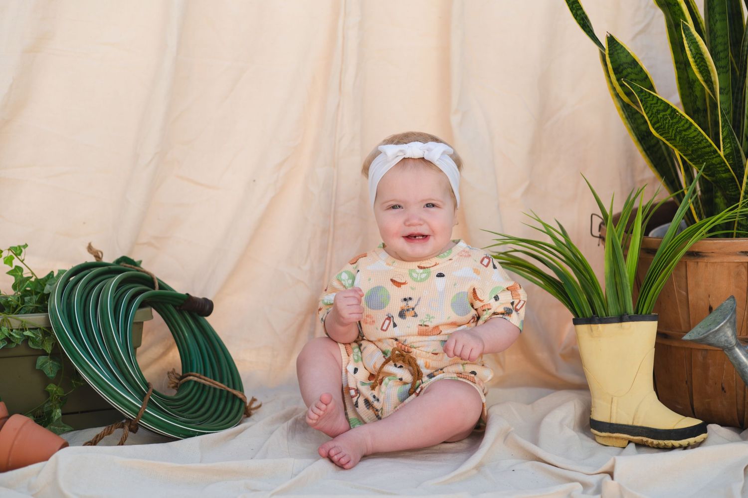 A baby in floral outfit sits among indoor plants and garden accessories.
