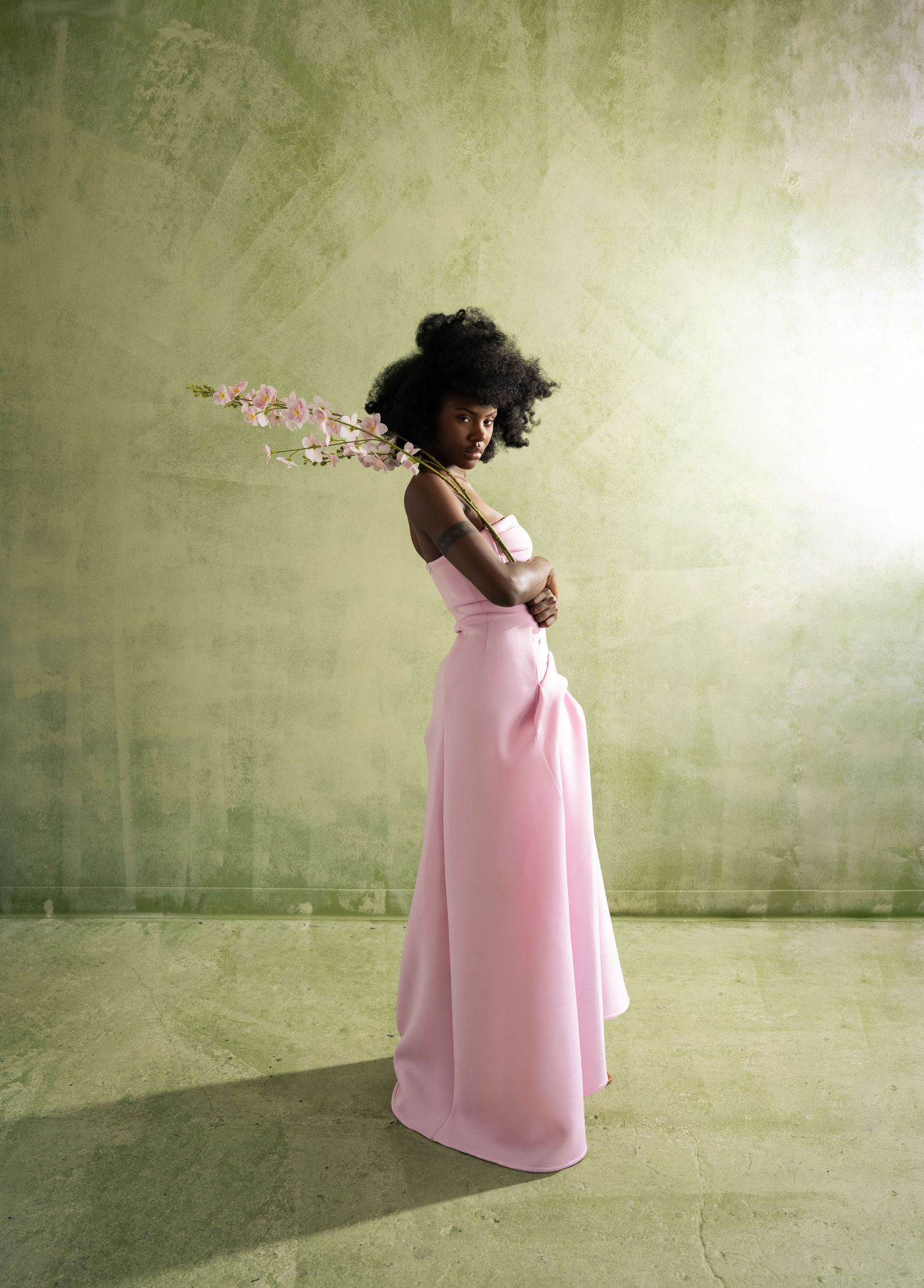 A black woman in a flowing pink dress twirling and dancing against a textured green wall.