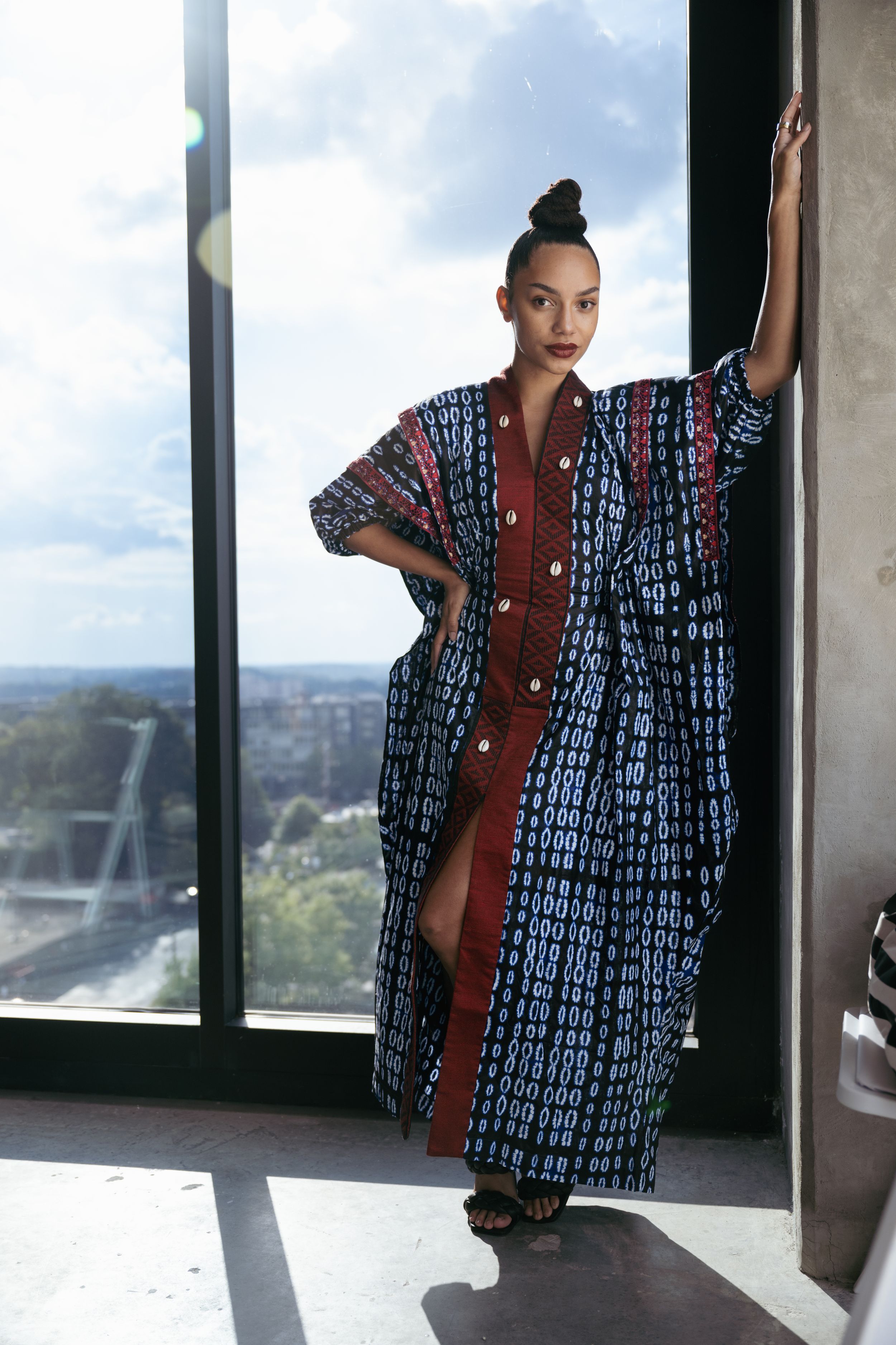 Model poses in a patterned blue and brown kimono-style dress against a cityscape window view.