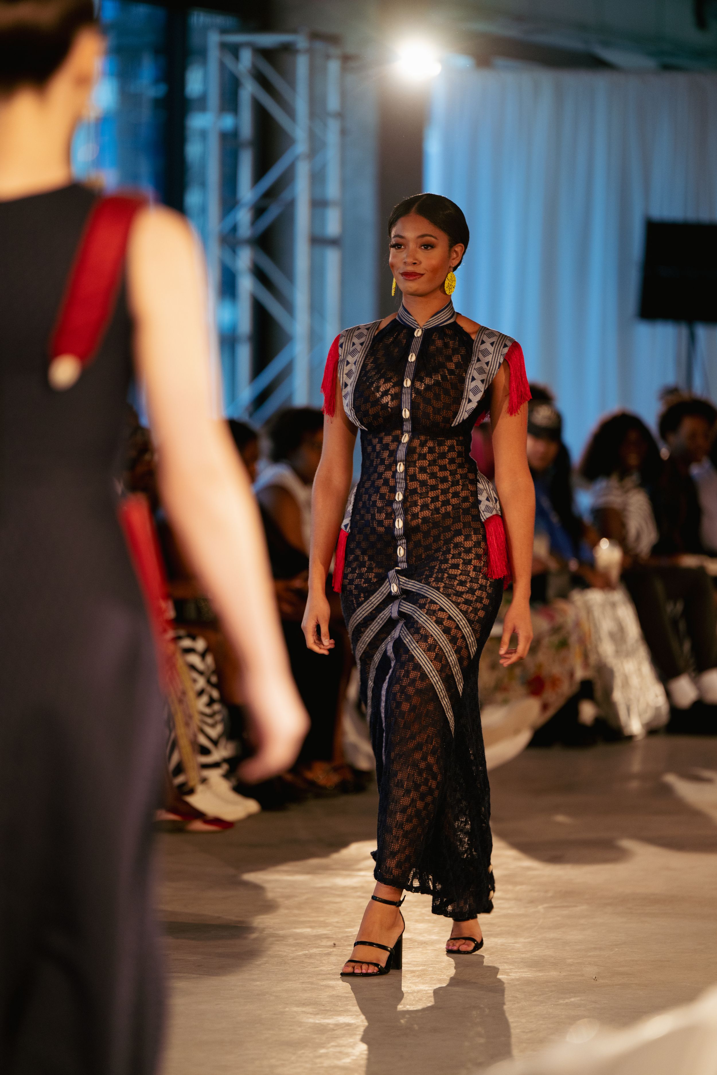 Model walks runway in sheer black patterned dress during fashion show.