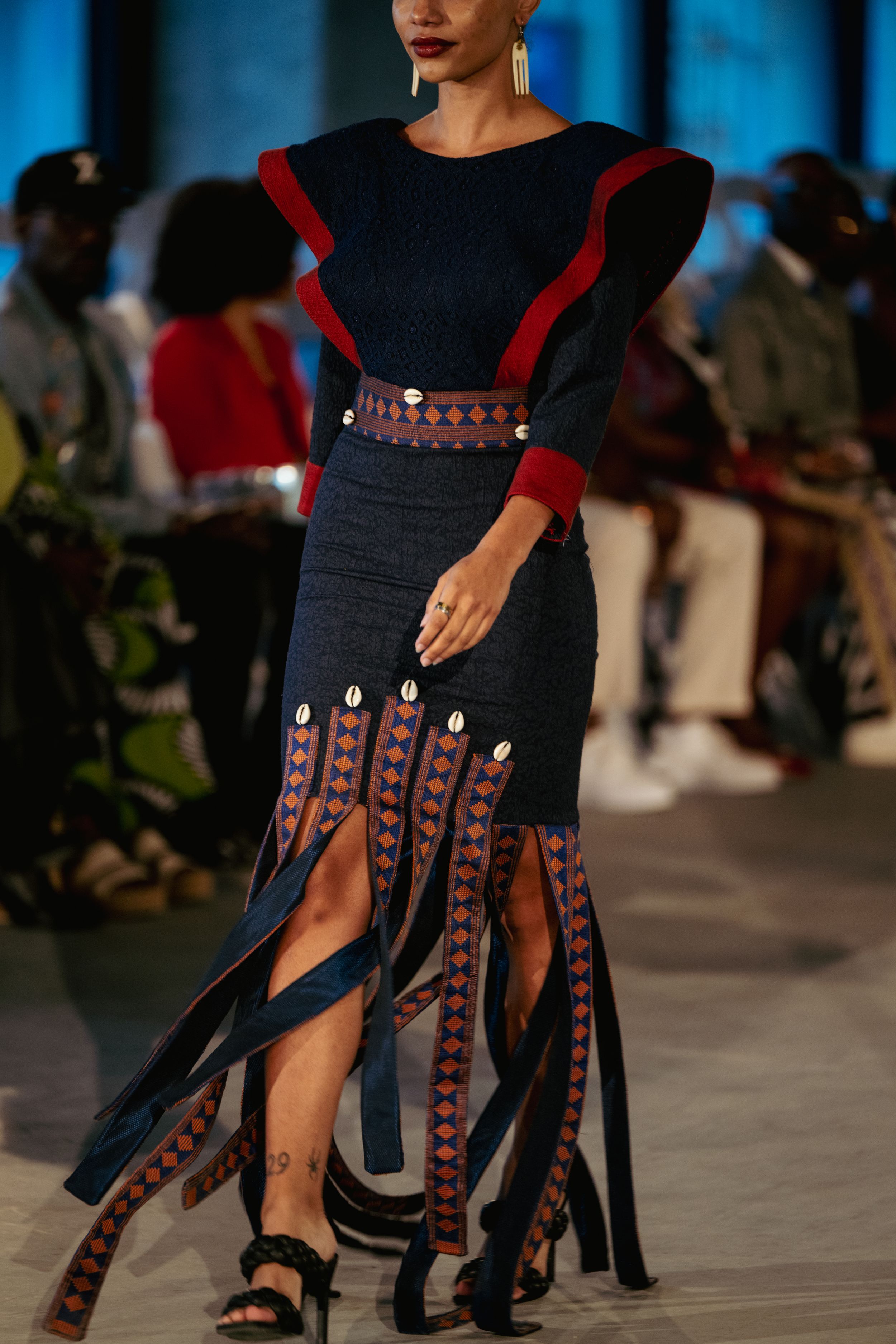 Navy dress with red trim and decorative cowrie shell details on fashion runway.