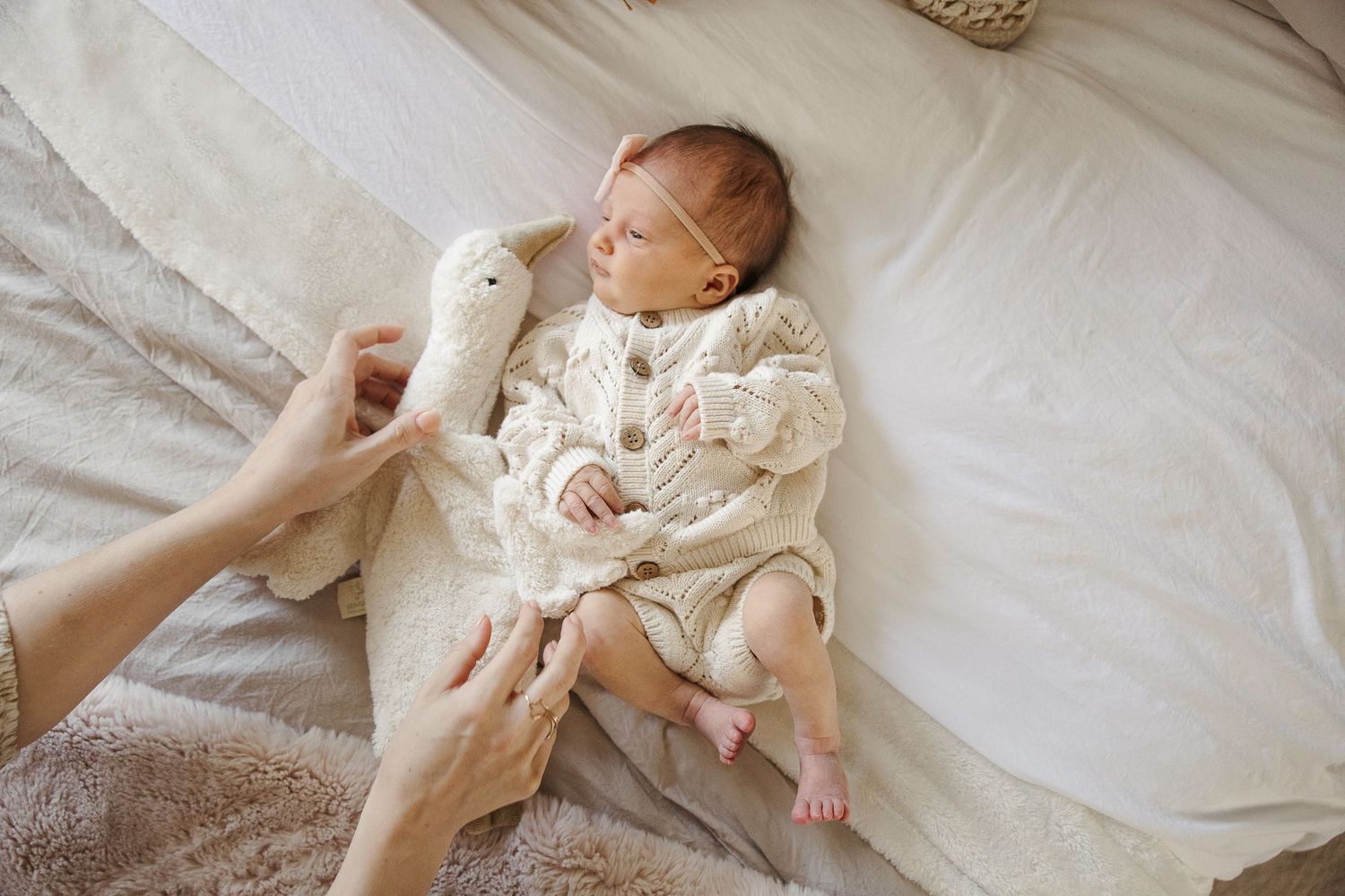 Baby wearing white knit sweater lies on bed while someone holds white stuffed bunny toy.