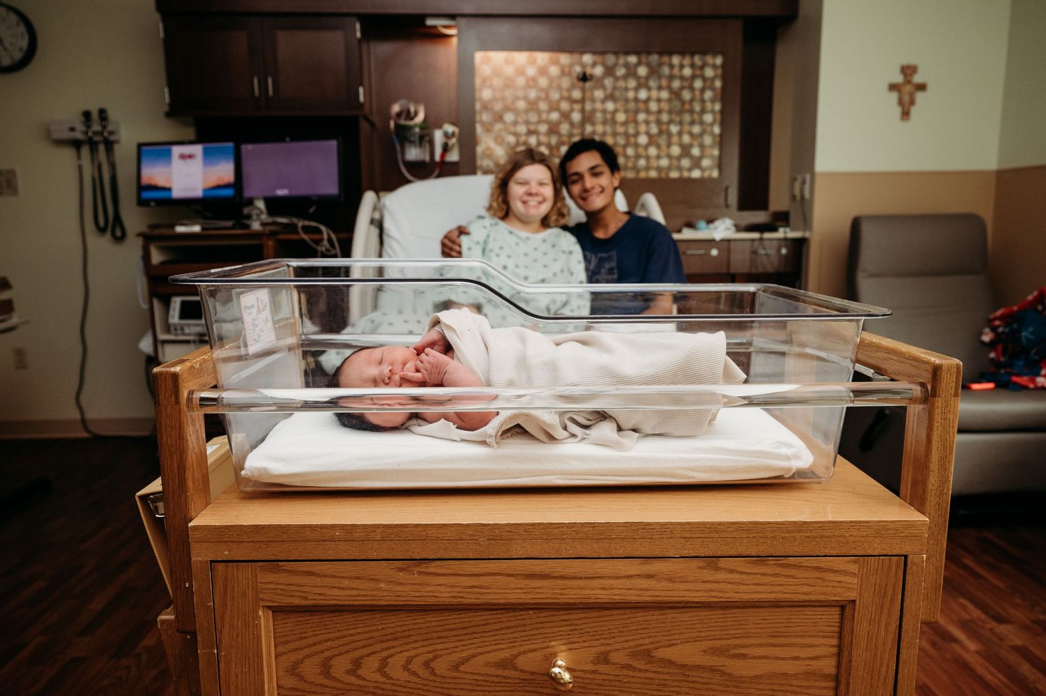 Mom and dad smile at their newborn in a bassinet in a hospital room.