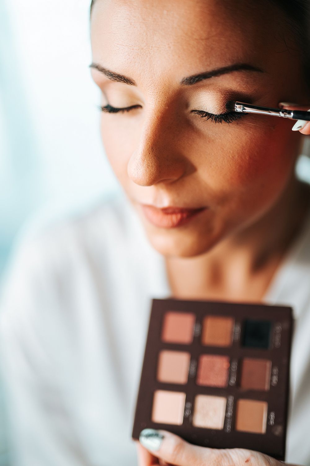 Bride applying eyeshadow with brush from brown palette during wedding makeup preparation.