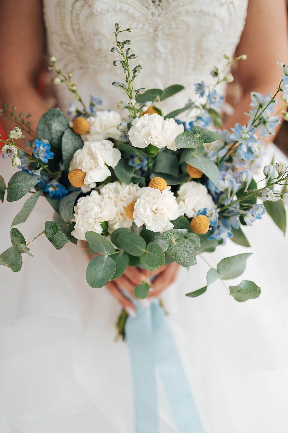 Bridal bouquet featuring white carnations, blue flowers, eucalyptus, and yellow billy balls with ribbon.