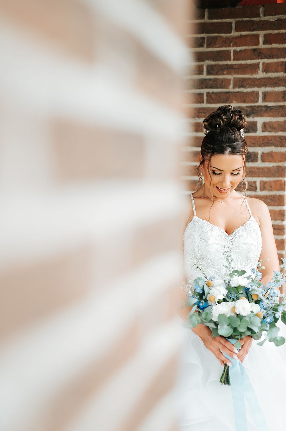 Bride in lace wedding gown holding blue and white bouquet against rustic brick wall.