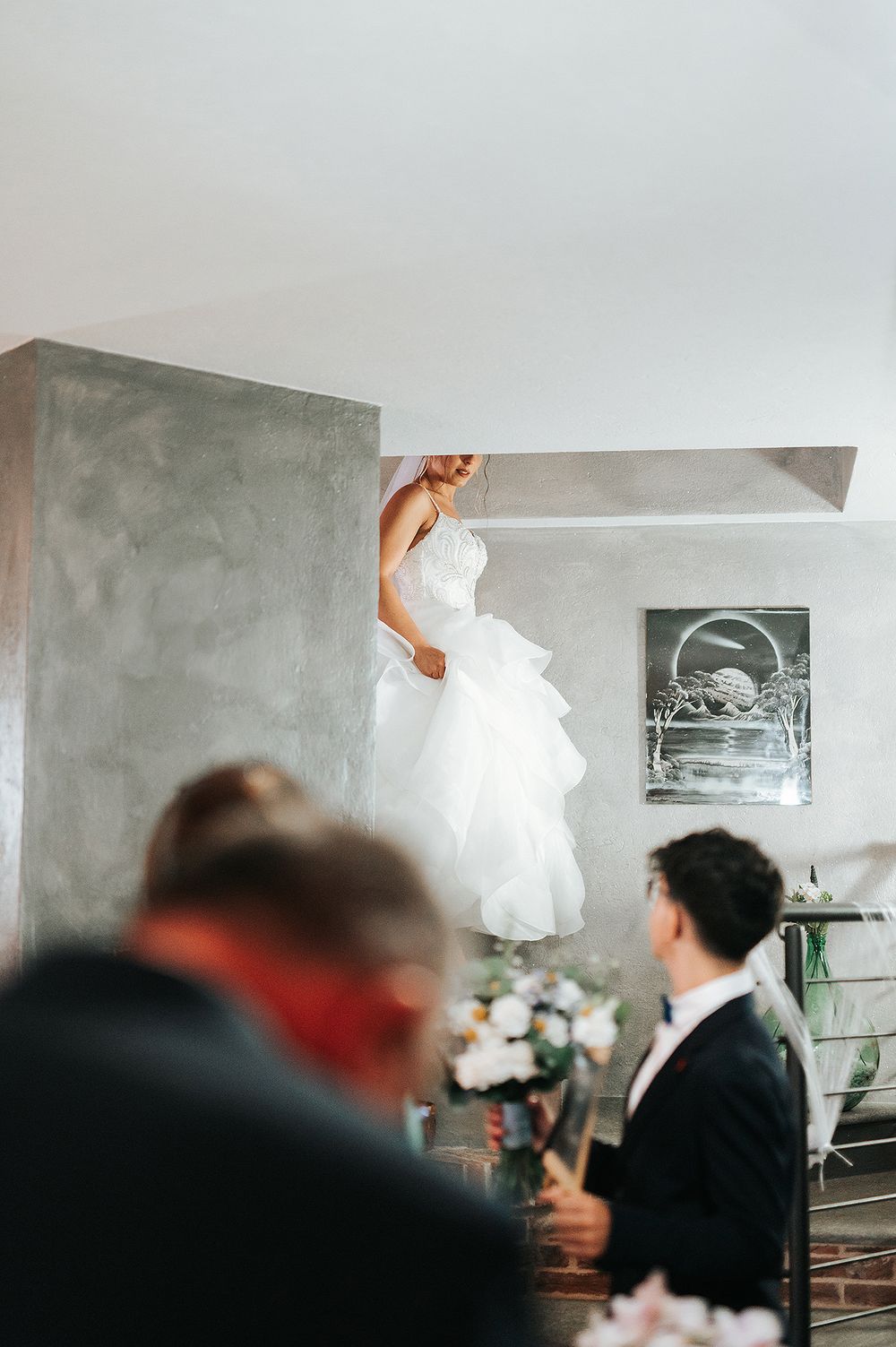 Bride in ruffled white gown descending stairs as groom watches during wedding first look.