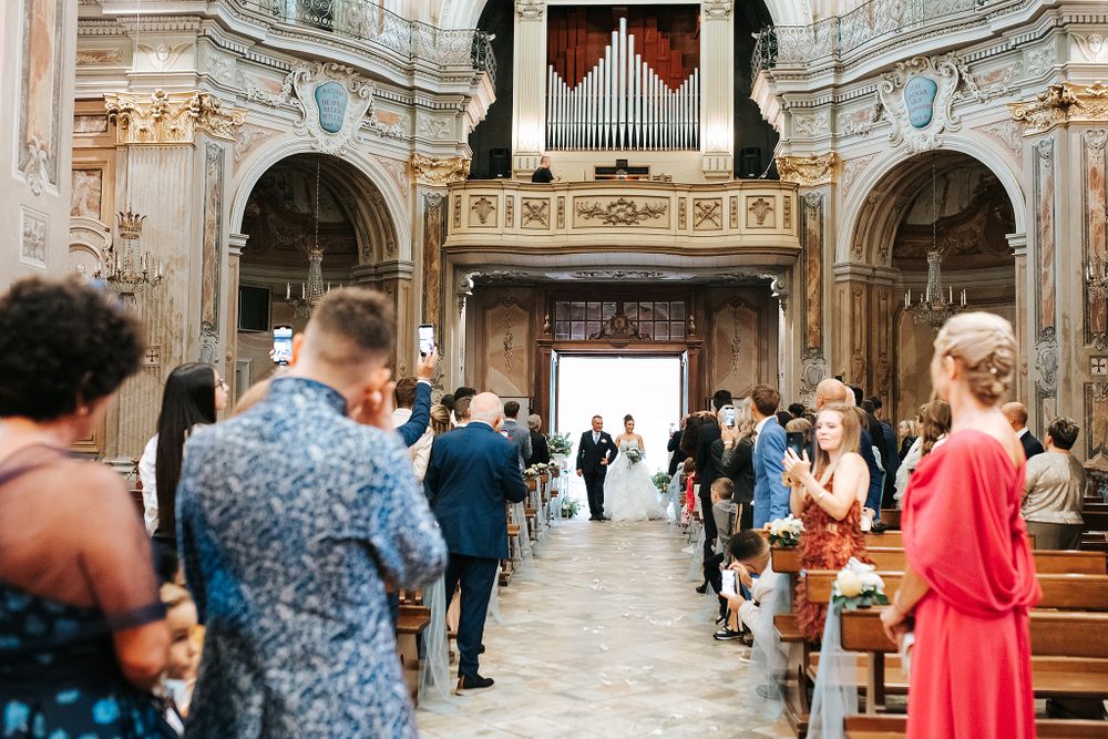 Wedding ceremony processional in ornate baroque church with guests standing in decorated wooden pews.