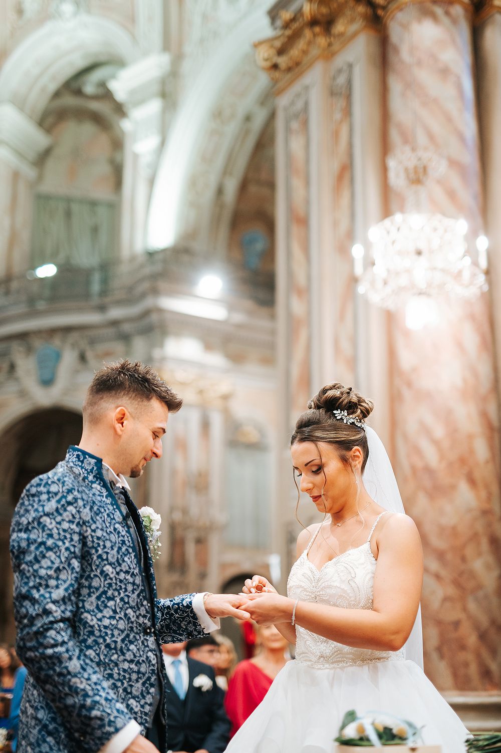 Bride places wedding ring on groom's finger during elegant church ceremony with chandelier backdrop.